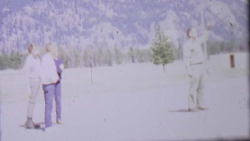 1967 8mm frame from Farragut State Park metadata showing two adult men standing together at left and another man moving at right, with a pale field and mountain forest behind them.