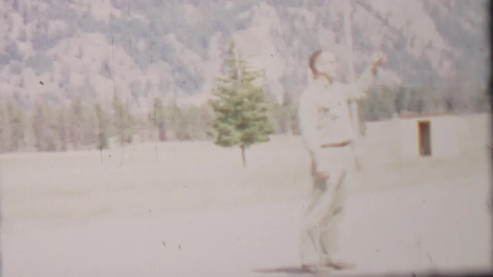 1967 8mm frame from Farragut State Park metadata showing an adult man in light clothing pointing upward in a grassy field, with evergreens and a mountain slope behind him.