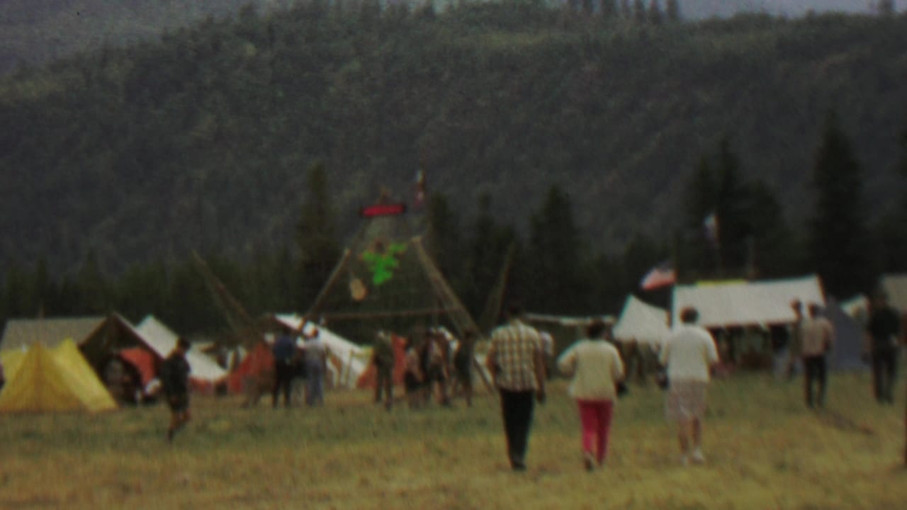 Wide view of a grassy festival field with tents, people, forested hills, and a wooden display frame.