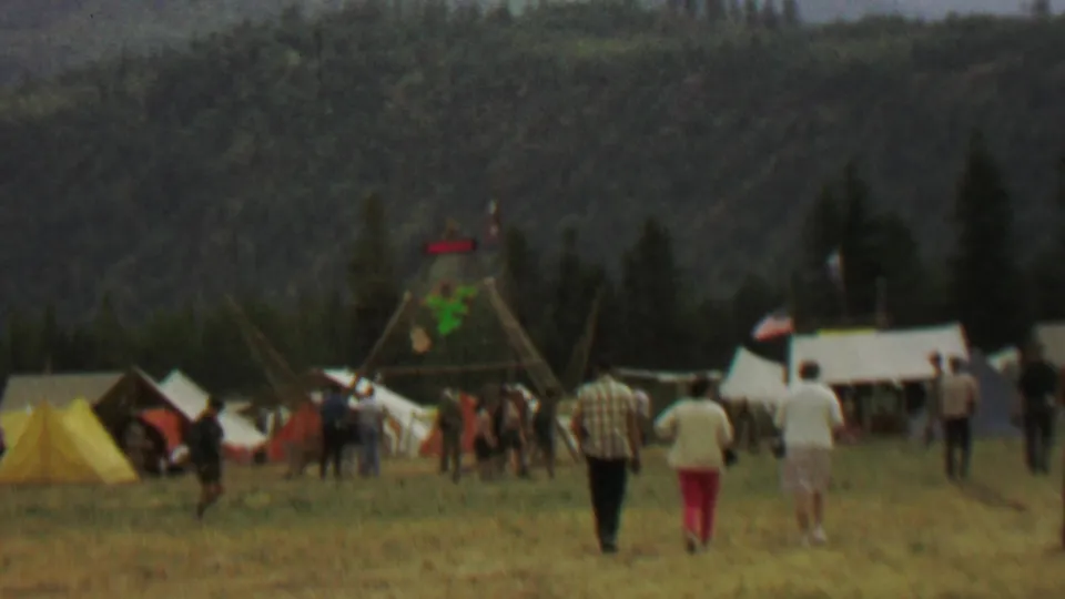 Wide 1967 8mm view at Farragut State Park showing people walking through a grassy festival setup area with tents, forested hills, and a wooden frame marked with a MEXICO sign.
