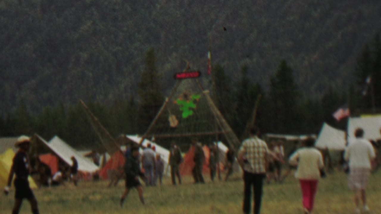 People walk past tents and a tall wooden frame with a MEXICO sign in a wooded field.