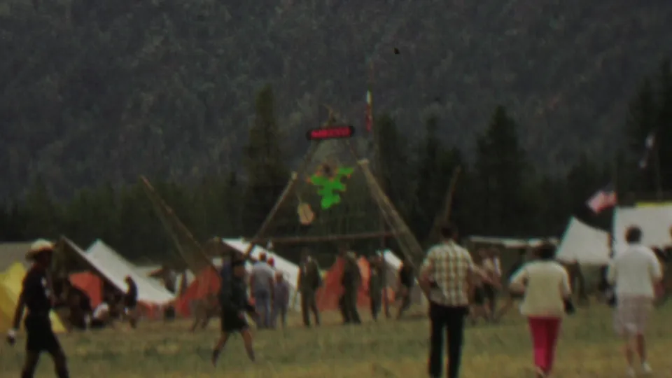 People in casual 1960s clothing move through a Farragut State Park festival setup area with white canvas tents, a tall wooden frame, a green cactus decoration, and a MEXICO sign.