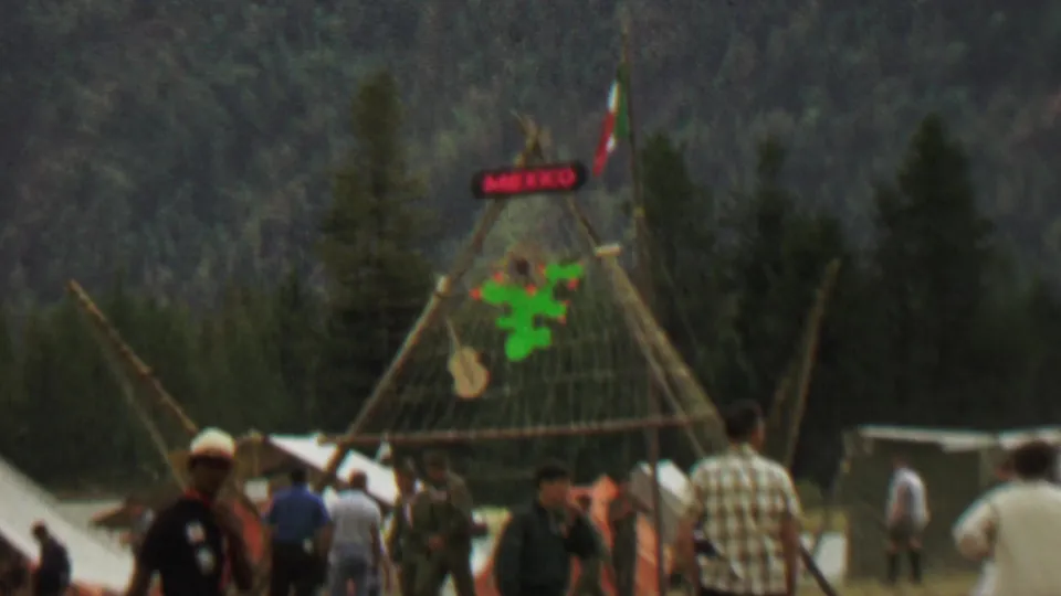 Closer 8mm frame of the festival display area, with a red MEXICO sign, green cactus decoration, Mexican flag, canvas tents, and attendees moving around the structure.