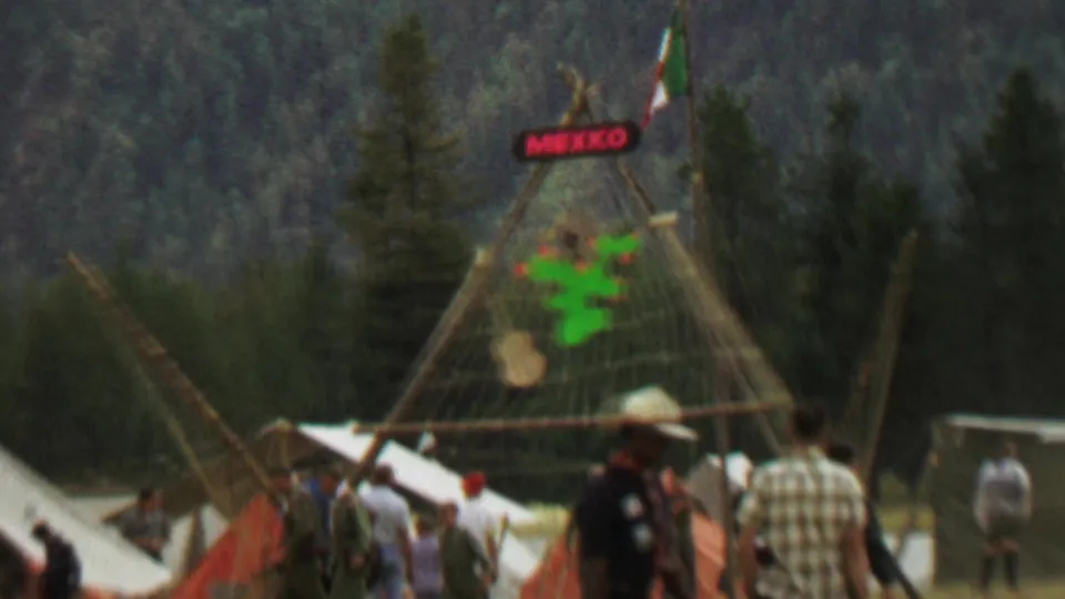 End frame from the 1967 Idaho home movie showing attendees near white canvas tents and a wooden MEXICO display structure, with forested hills behind the festival setup.