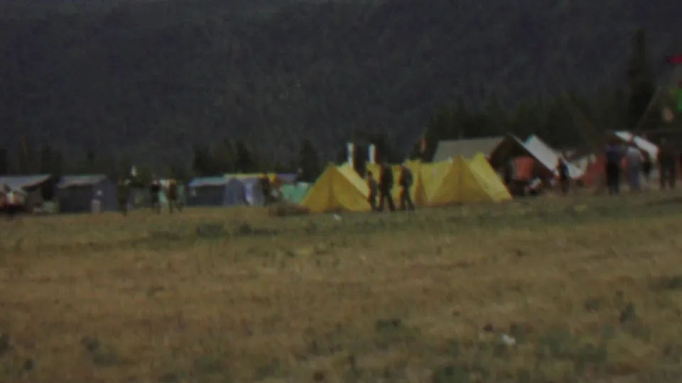 Wide 1967 park festival view with yellow tents, scattered visitors, a Canadian flag, and a wooded hillside beyond the open grass.
