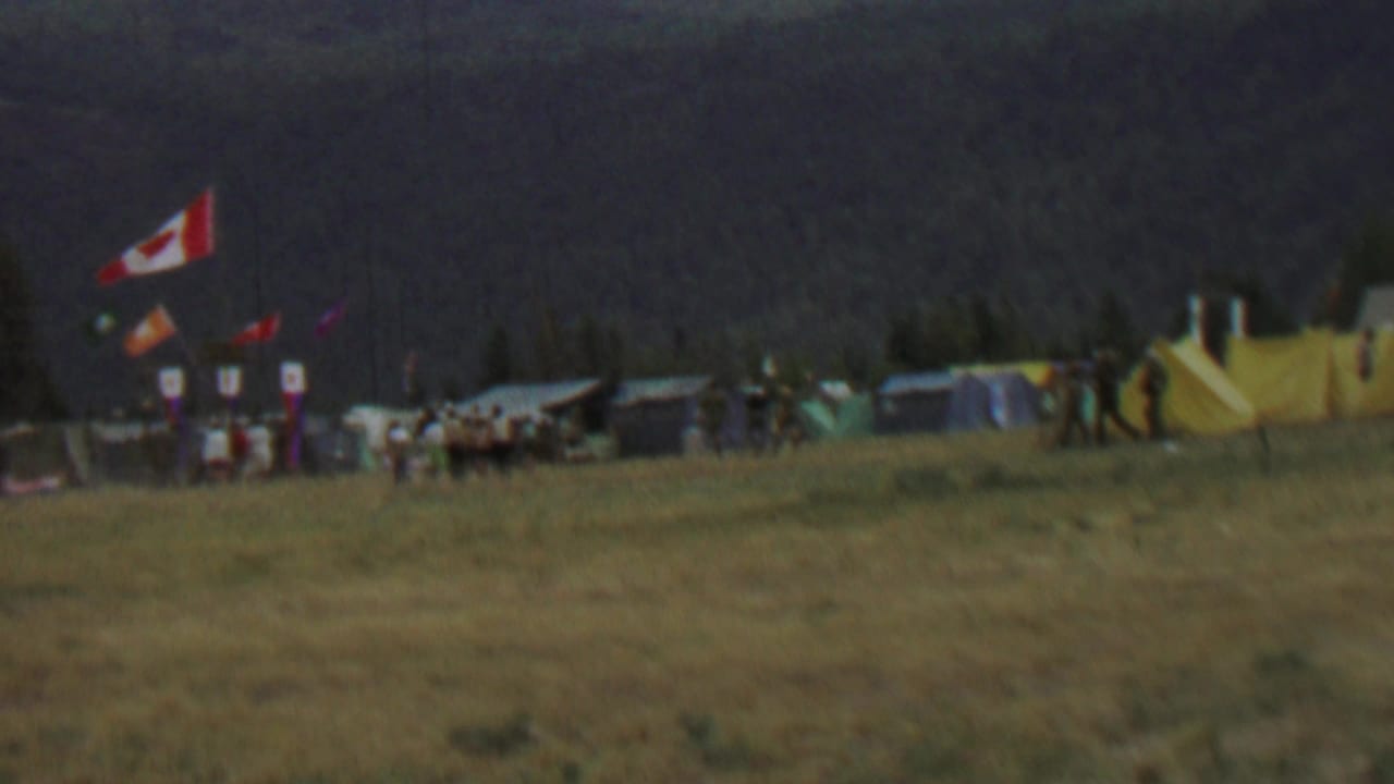 1967 8mm view at Farragut State Park, Idaho, showing a distant scout campsite with a Canadian flag, tents, smaller banners, and people moving across a grassy field.