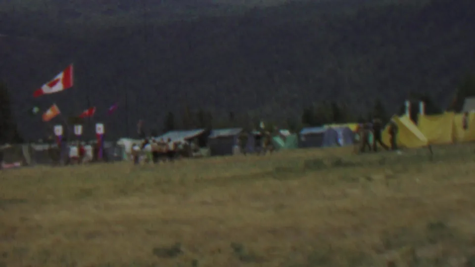 1967 8mm view at Farragut State Park, Idaho, showing a distant scout campsite with a Canadian flag, tents, smaller banners, and people moving across a grassy field.