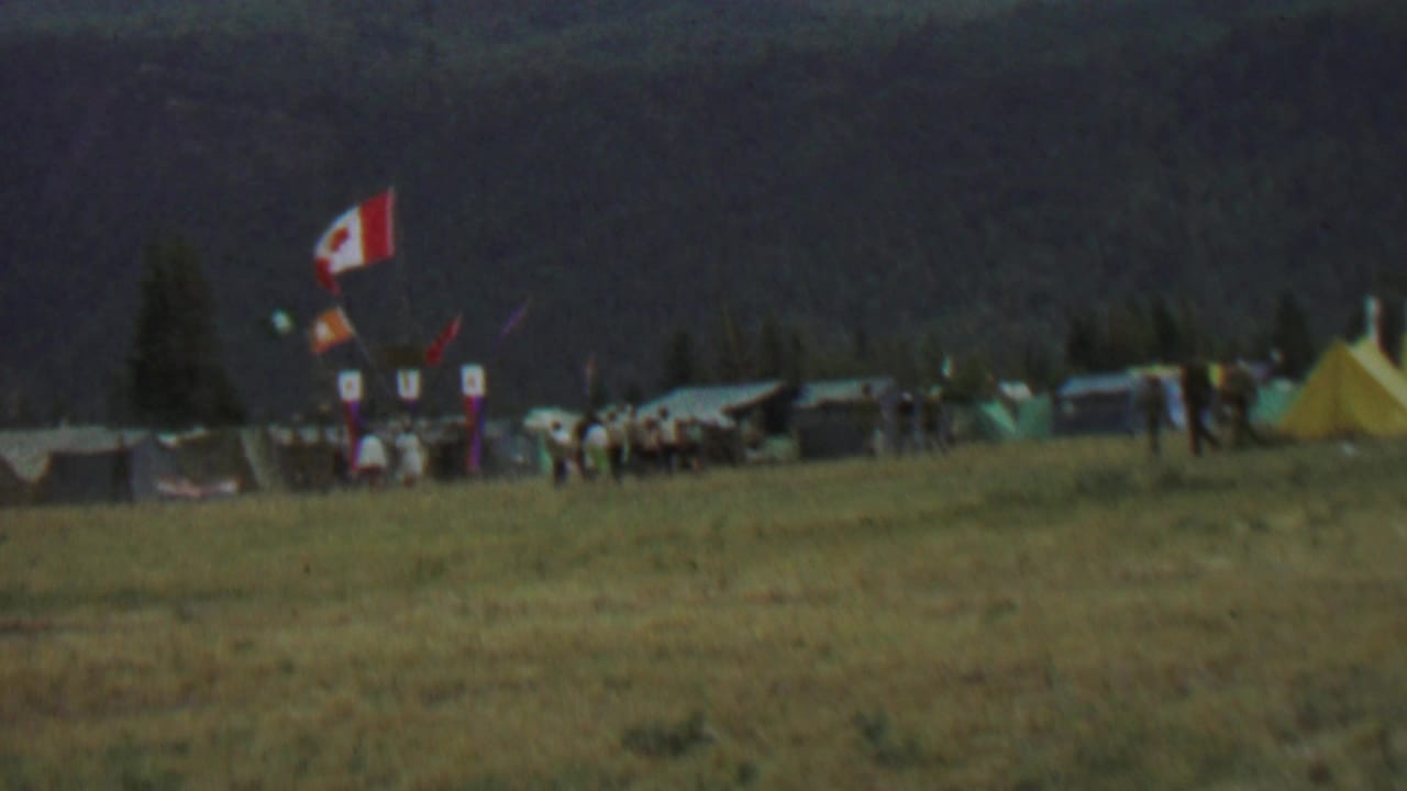 Campsite with Canadian flag, tents, and people gathered near banners below a wooded hillside.