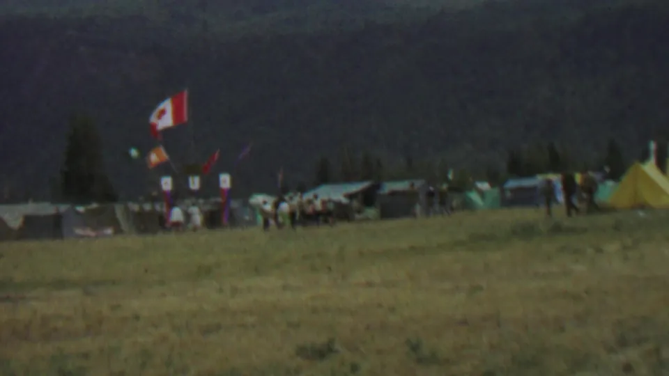 Distant 1967 home-movie frame from Farragut State Park showing a Canadian flag above tents, banners, and people gathered in a field beneath a dark wooded hillside.