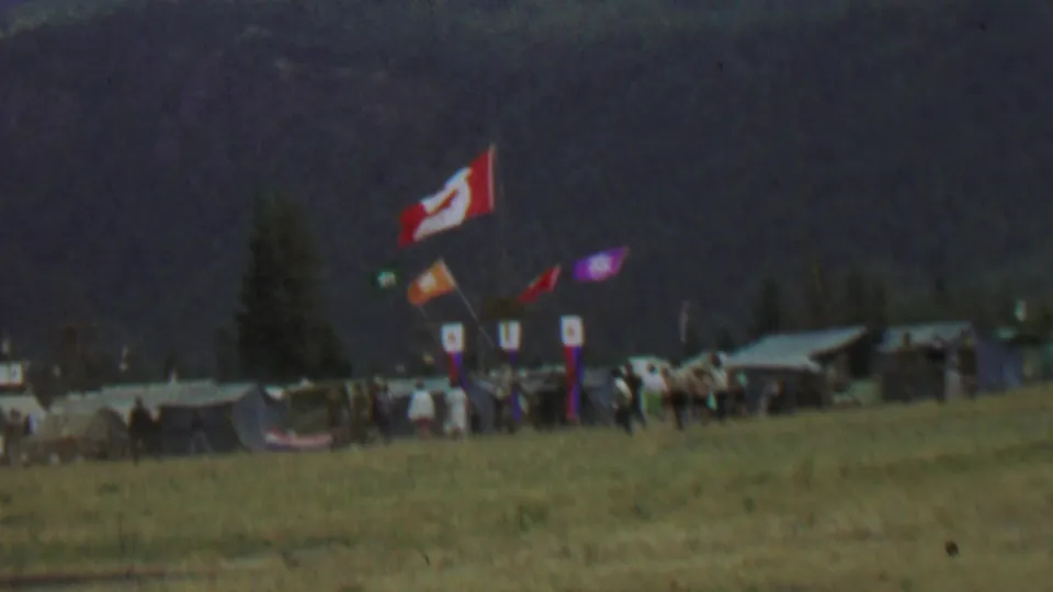 1967 8mm frame at Farragut State Park showing a flag cluster, tents, and people standing or walking near a scout campsite in a grassy field.