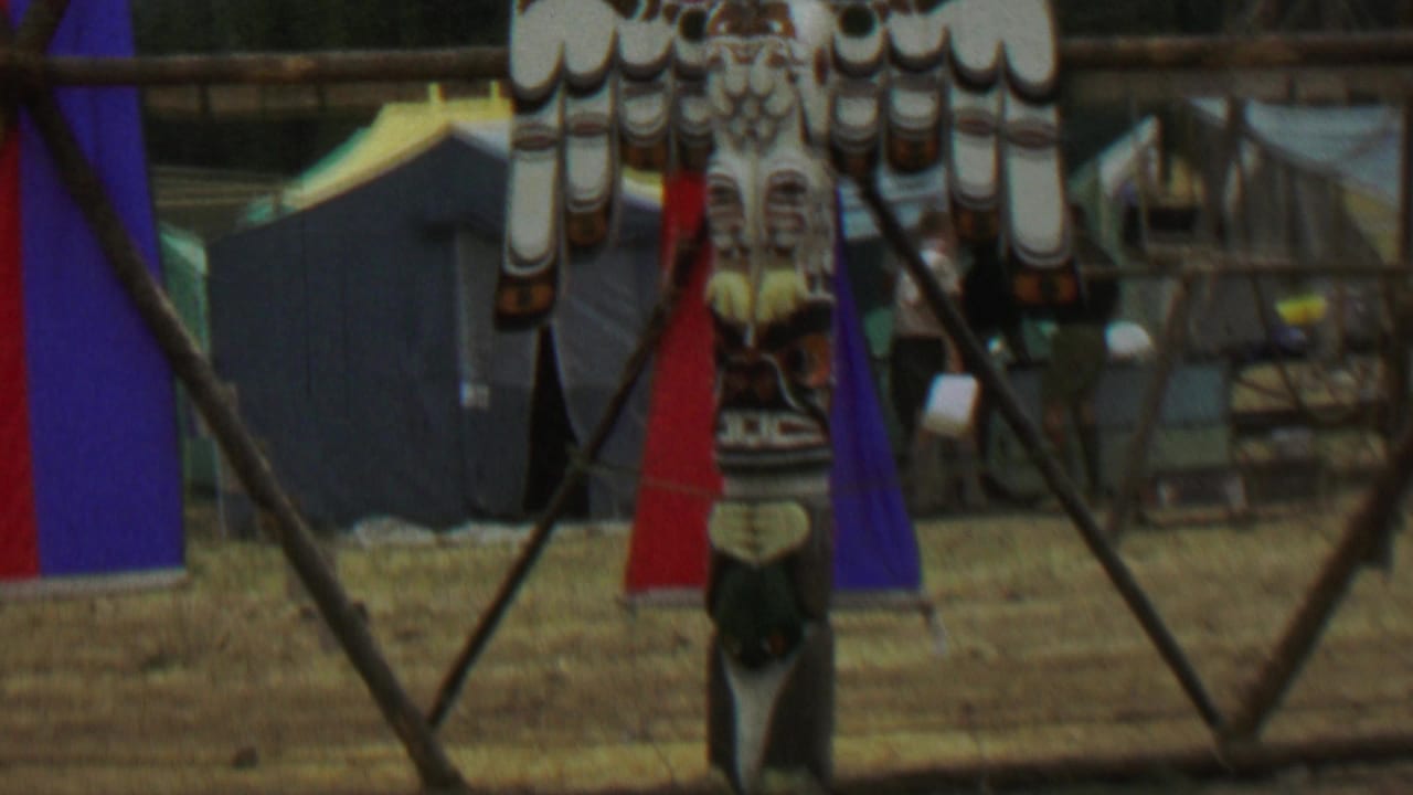 1967 8mm frame from Farragut State Park showing a tall carved totem pole with a prominent bird design, set among tents, wooden supports, and red and blue fabric banners.