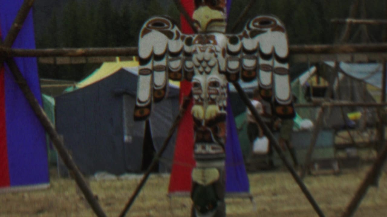 1967 Farragut State Park frame with the painted bird-topped totem pole centered against a campsite backdrop of tents, fabric banners, wooden rails, and distant people.
