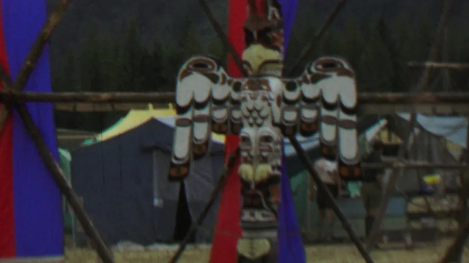 Soft 8mm frame showing a closer view of the carved bird design on the totem pole, with red and blue fabric, tents, and wooden structures behind it at the Idaho park gathering.