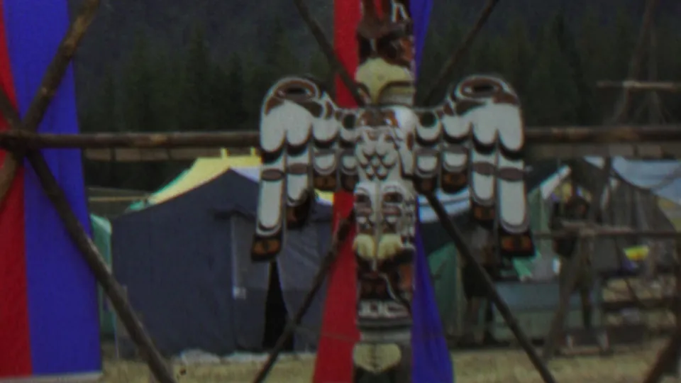 Final 1967 frame of the totem pole display at Farragut State Park, with the bird carving, colored fabric banners, tents, wooden supports, and distant people visible in the background.