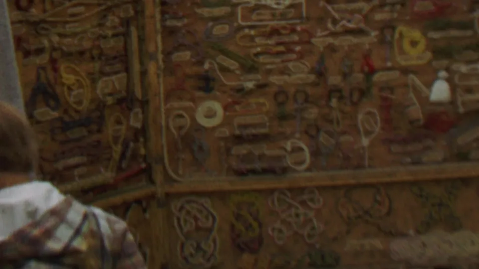 The final frame shifts to a close, soft-focus view of a decorated wooden panel with carved or painted symbols, with only partial bodies or objects at the edge of the frame.