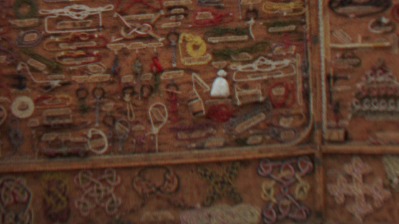 1967 United States 8mm frame showing a warm-toned handmade craft display with embroidered shapes, trinkets, and jewelry-like objects arranged in a grid.