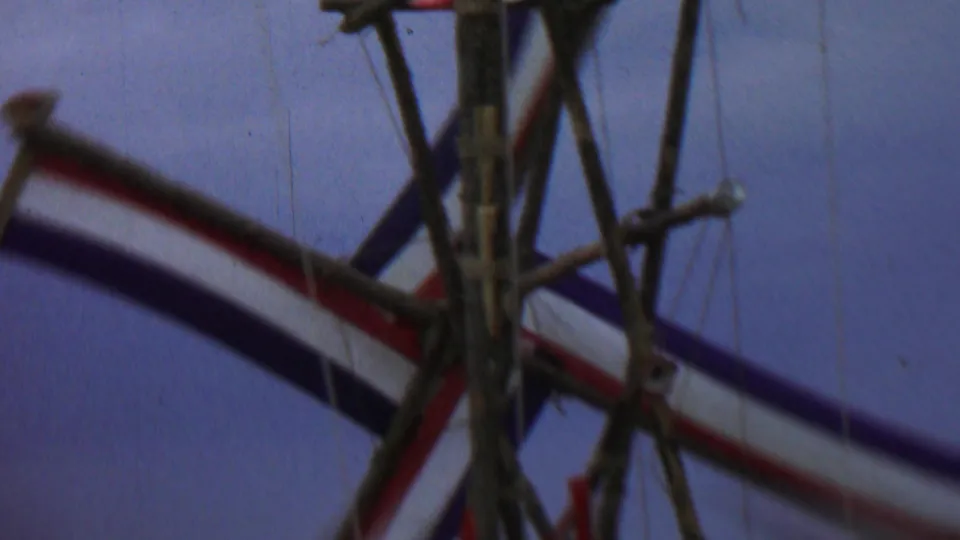 Close 1967 Idaho home-movie view of a weathered wooden windmill-like structure with red, white, and blue striped fabric set against a purple-blue sky.
