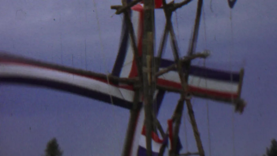Soft 1967 8mm frame of the rustic windmill-like structure, with crossed wooden supports and patriotic-colored fabric moving through the composition.