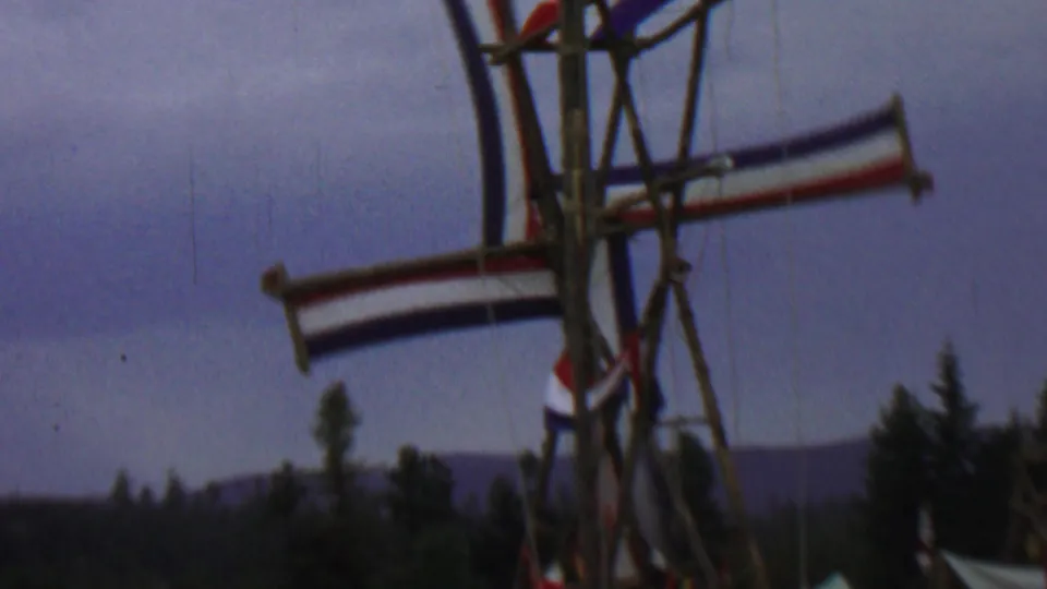 Wider 1967 Idaho 8mm view of the rustic windmill-like structure with red, white, and blue fabric, a dark tree line, and distant hills under a dusky sky.