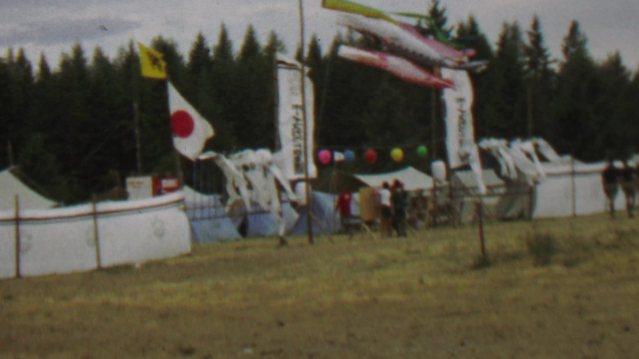 1967 8mm frame from Farragut State Park showing a windy festival entrance with a Japanese flag, carp streamers, tents, balloons, visitors, dry grass, and dark evergreen forest behind.