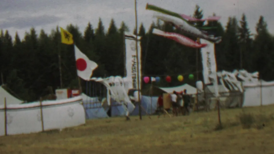 A 1967 home-movie frame shows a windy festival area at Farragut State Park with white tents, carp streamers overhead, flags, colored balloons, a small group of visitors, dry grass, and evergreen trees.
