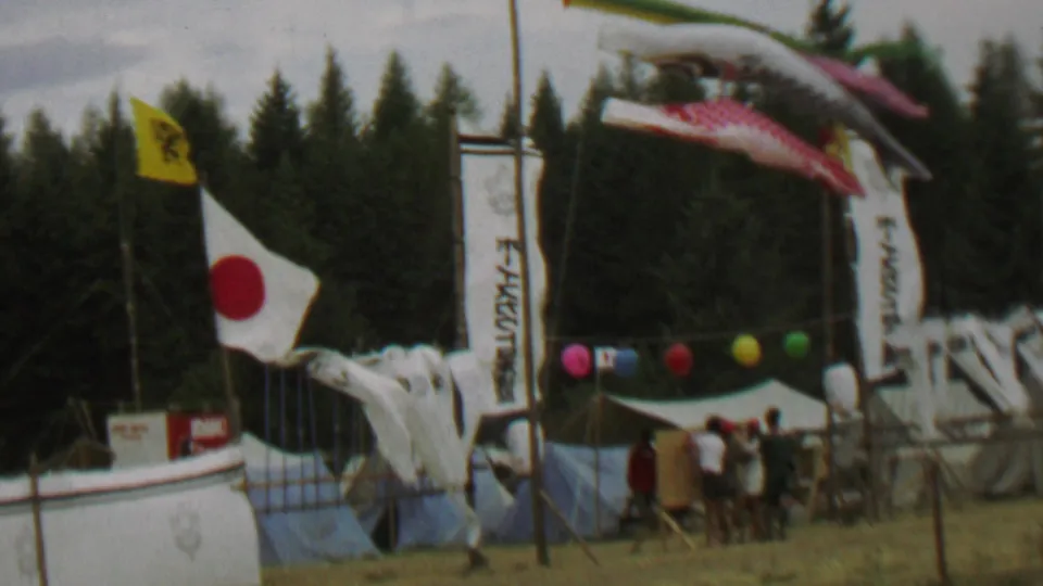 Closer 1967 Farragut State Park festival frame with a Japanese flag in the wind, a large carp streamer overhead, vertical banners, colored balloons, people at the entrance, and a dense evergreen backdrop.