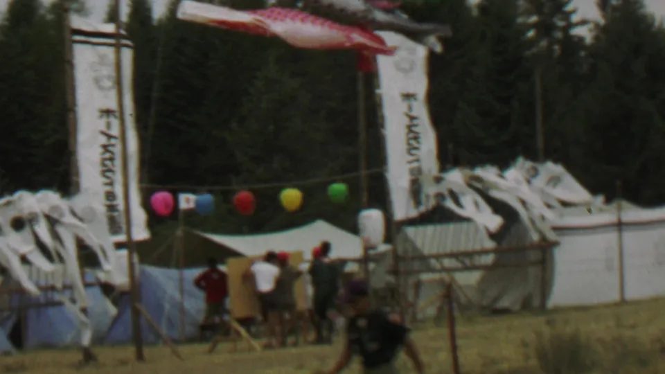 A wider 1967 8mm view of the Japanese festival area shows carp streamers, banners with Japanese text, balloons, tents, and visitors standing near a fence.