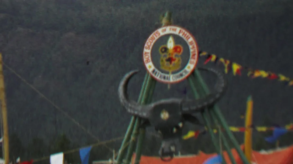 1967 home-movie frame from Farragut State Park showing the bull's head attraction beneath the Boy Scouts of the Philippines emblem, with red, yellow, blue, and white pennants and a tall flagpole nearby.