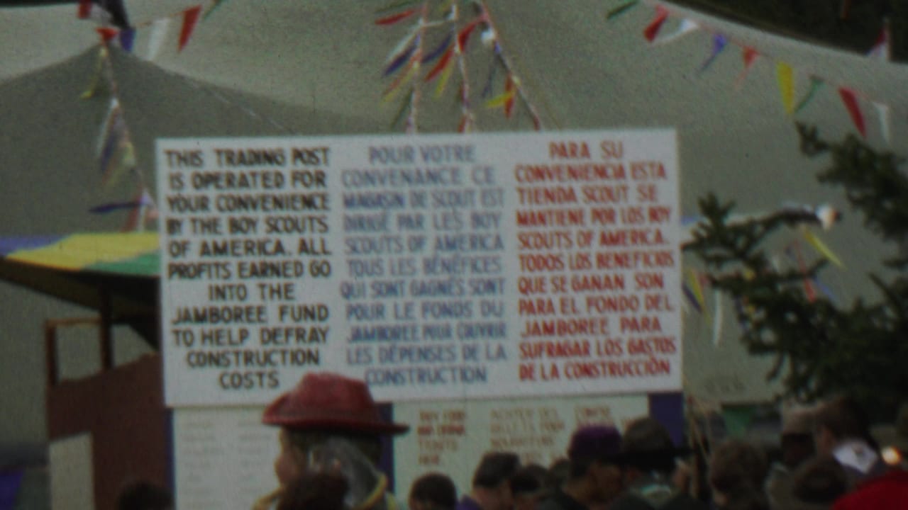 1967 Farragut State Park 8mm frame showing a large English, French, and Spanish Boy Scouts trading post sign with pennants and attendees below.