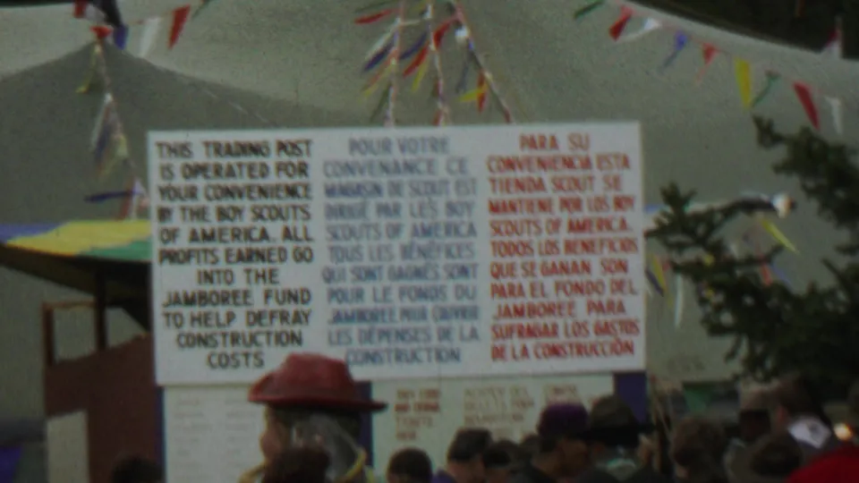 1967 Farragut State Park 8mm frame showing a large English, French, and Spanish Boy Scouts trading post sign with pennants and attendees below.