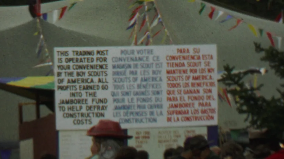 Large Boy Scouts trading post sign in English, French, and Spanish at the outdoor Farragut State Park gathering, with pennants and people visible below.