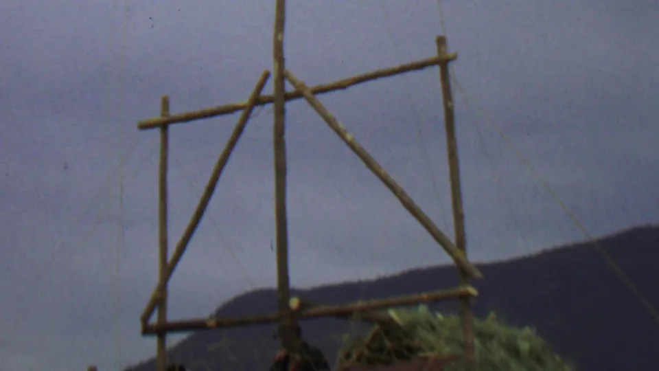 1967 home-movie frame of a rustic wooden pole frame with a partly visible hanging flag or fabric and distant hills under a cloudy sky.