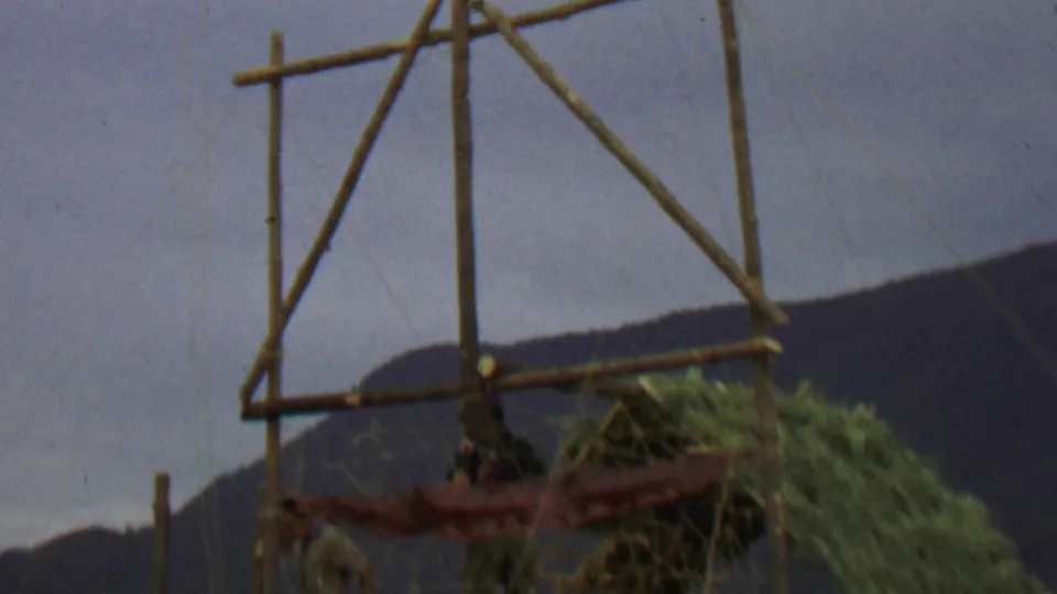 1967 8mm frame showing a rustic wooden structure with a red hanging fabric or flag, people partly visible beneath it, and hills in the background.