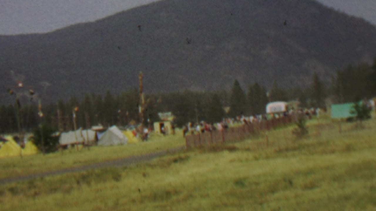 1967 8mm view of a grassy field at Farragut State Park with small tents, distant people, flags, trees, and mountains in the background.