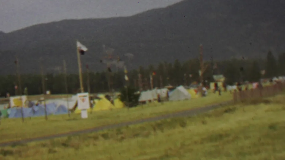 Handheld 8mm footage shows distant people near tents and a flagpole in a grassy Farragut State Park field, with dark mountains behind the tree line.