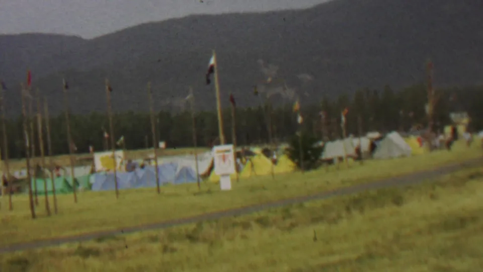 The final frame shows poles and small flags in the foreground, tents and people across the field, and the wooded mountain landscape of Farragut State Park behind them.