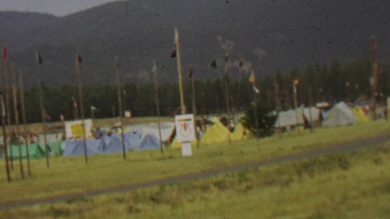 1967 home-movie view at Farragut State Park showing a grassy tent camp with blue and yellow tents, tall flagged poles, posted signs, trees, and mountains in the background.