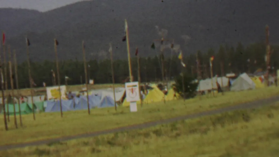 1967 home-movie view at Farragut State Park showing a grassy tent camp with blue and yellow tents, tall flagged poles, posted signs, trees, and mountains in the background.