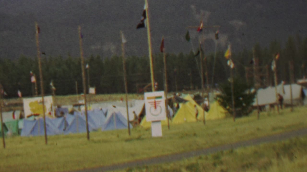 Tent camp with blue and yellow tents, flags, signs, grass, trees, and mountains.