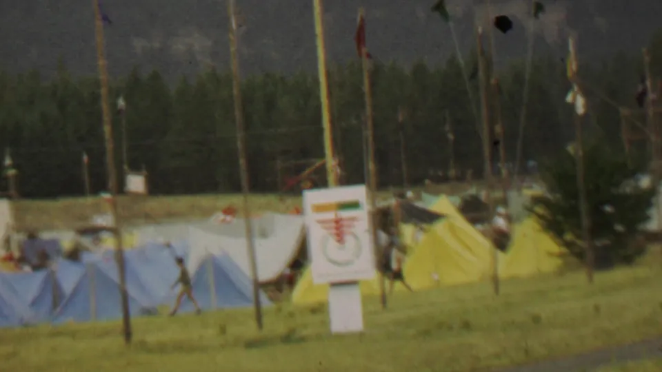 Closer 1967 campsite view showing blue, yellow, green, and white tents, tall flagpoles, a posted sign, wooded background, and a distant person within the camp area.