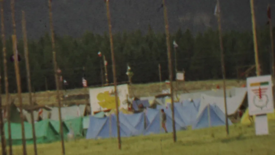 1967 Farragut State Park home-movie frame with blue tents, yellow sign or panel, tall flagpoles, wooded background, and a person walking near the camp structures.