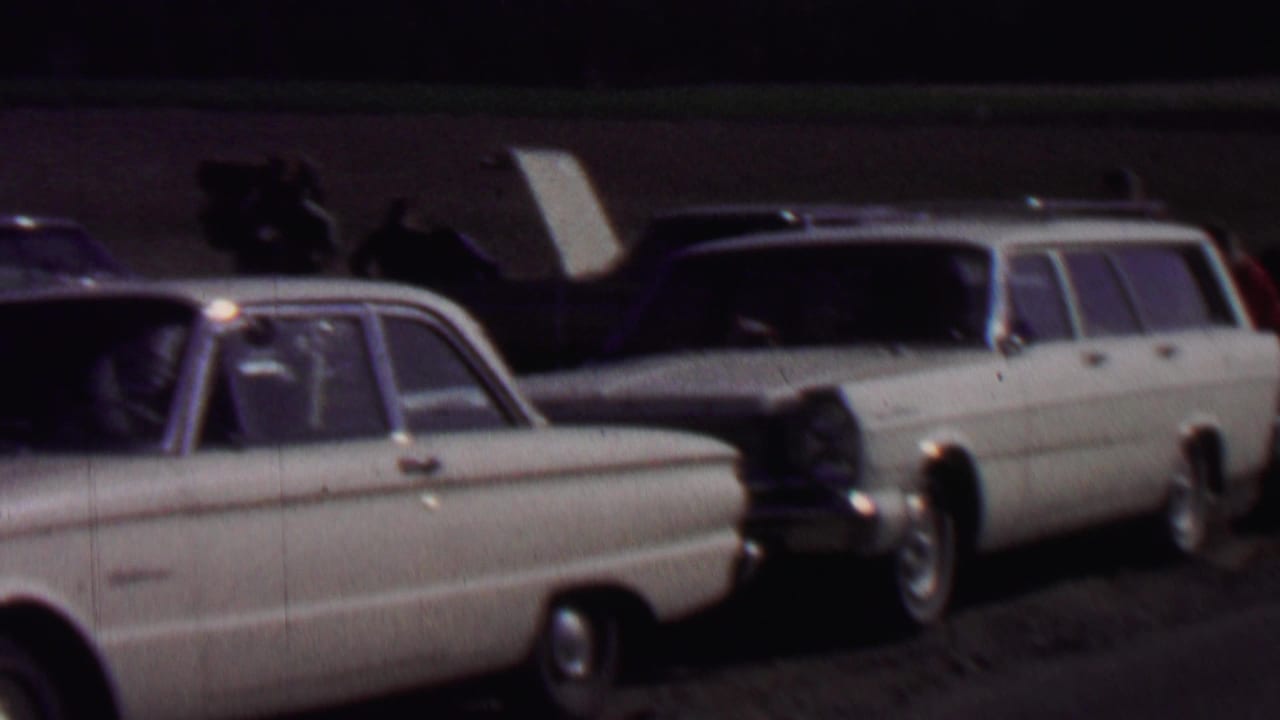 1967 home-movie frame from Farragut State Park, Idaho showing light-colored classic cars and a white station wagon with its rear hatch raised in a dim outdoor parking area.