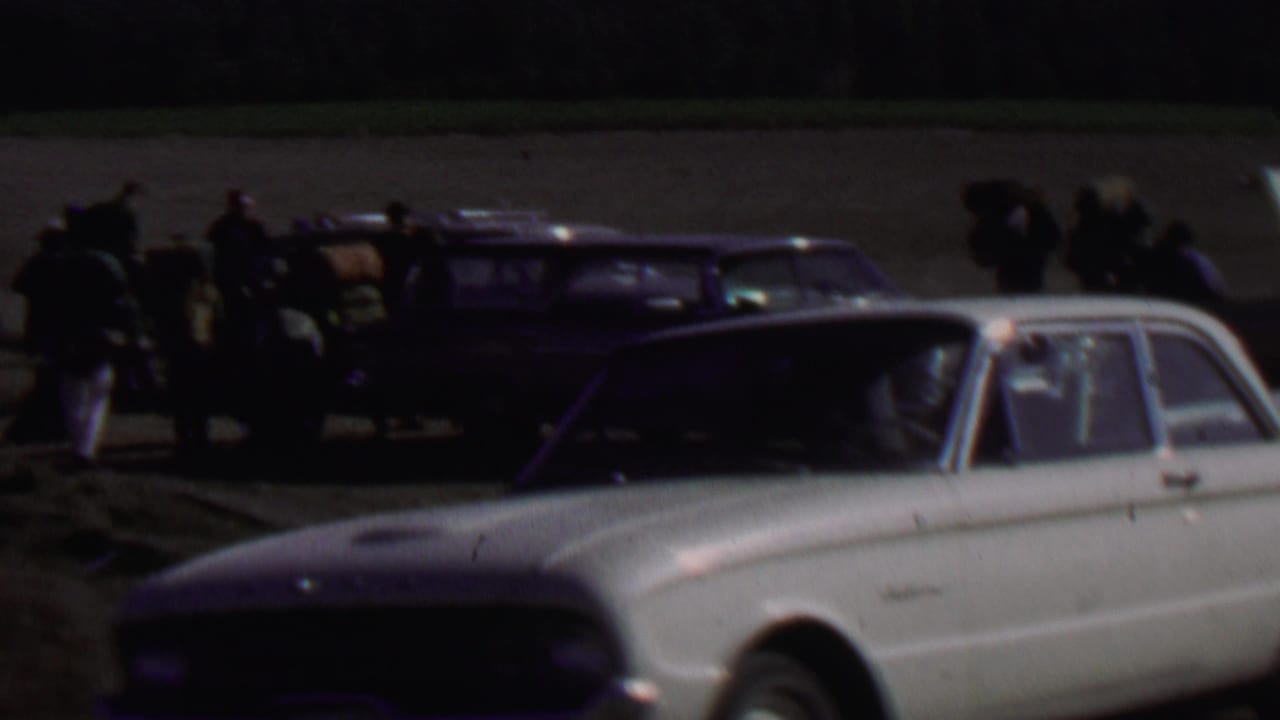 Distant 1967 Farragut State Park home-movie frame with a group loading or gathering near station wagons as a white compact car fills the foreground.