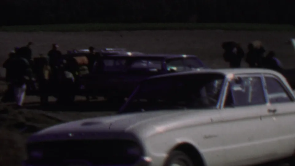 Distant 1967 Farragut State Park home-movie frame with a group loading or gathering near station wagons as a white compact car fills the foreground.