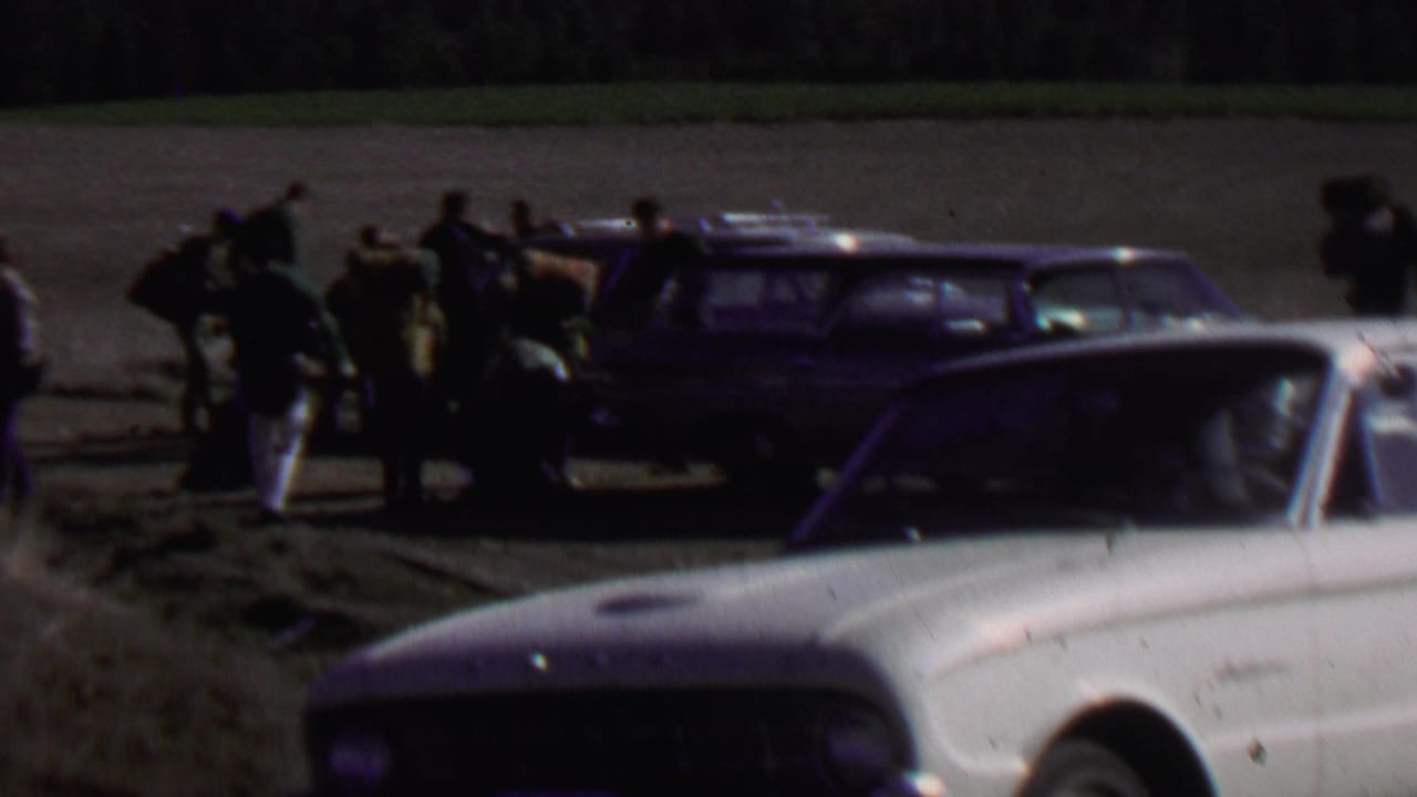 Group of people stand around a station wagon with gear beside parked cars.