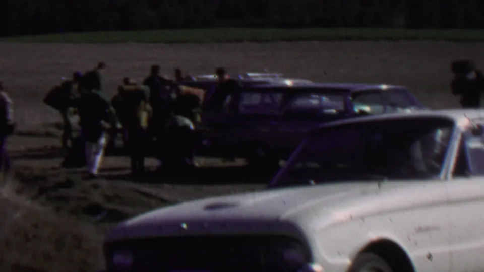 People in outdoor clothing gather around a dark station wagon and camping gear in a rural field setting, with another car partly visible in front.