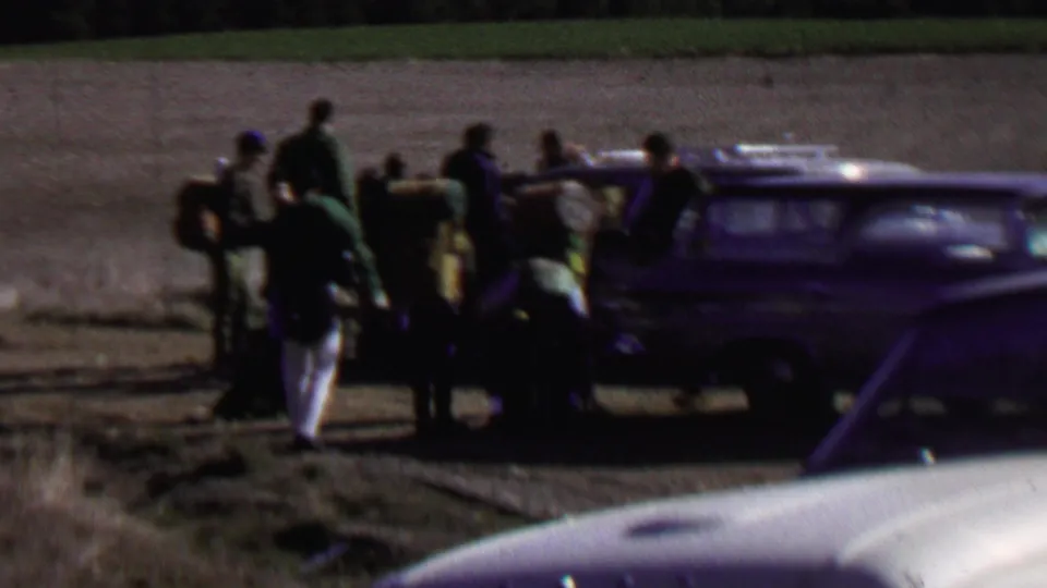 Distant 8mm frame of people handling backpacks and camping equipment near a dark station wagon at Farragut State Park, Idaho, based on supplied metadata.