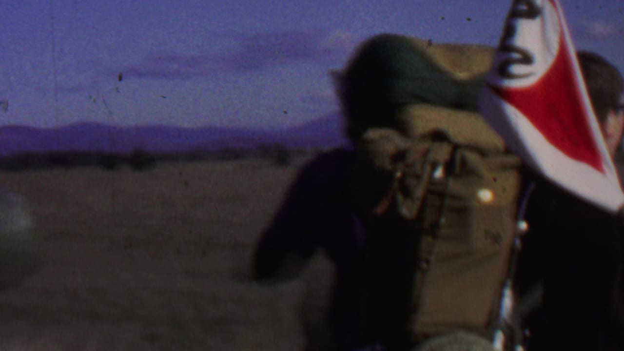 1967 8mm home-movie frame from Farragut State Park, Idaho, showing backpacked walkers from behind as they cross an open field with red and white flags.
