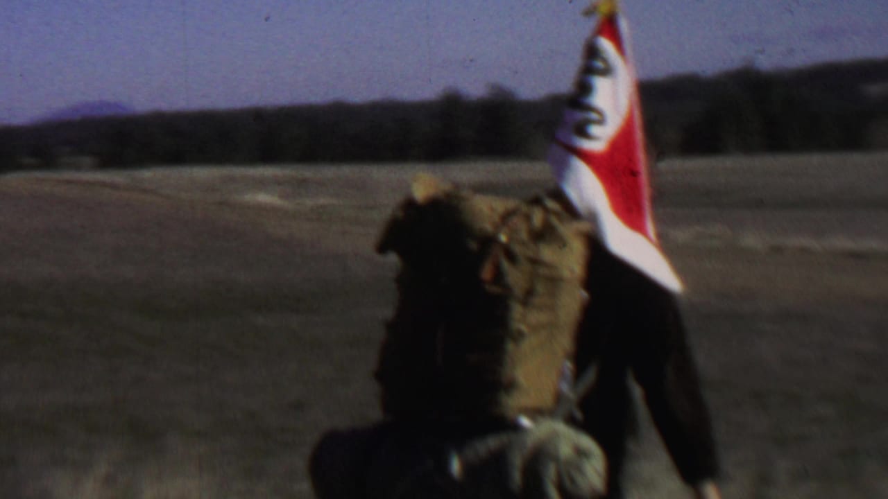 Backpackers continue across a field with flags and large packs visible from behind.