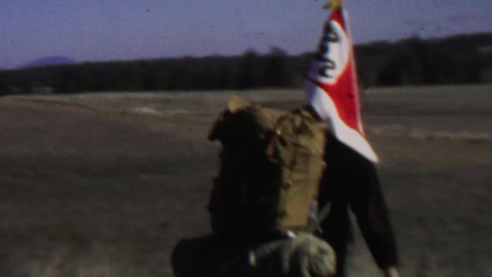 Backpackers in a 1967 Farragut State Park home movie move away from the camera across open ground, with flags, packs, hills, and sky visible.